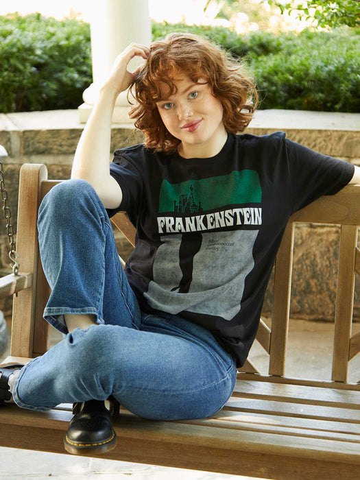 Person wearing a 'Frankenstein' t-shirt sitting on a wooden bench outdoors.