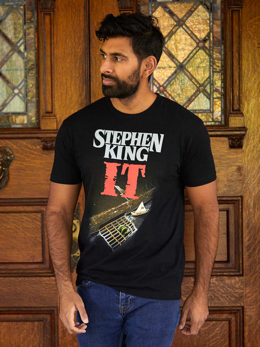 Man wearing a black t-shirt with 'Stephen King It' design in front of wooden paneling.
