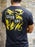 Person wearing a black t-shirt with yellow dragons and 'Earn One' text on the back, standing against a stone wall.