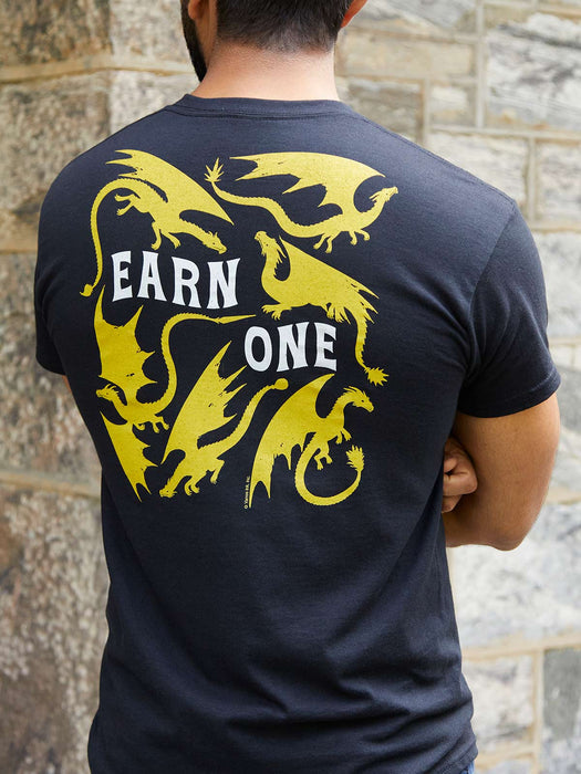 Person wearing a black t-shirt with yellow dragons and 'Earn One' text on the back, standing against a stone wall.