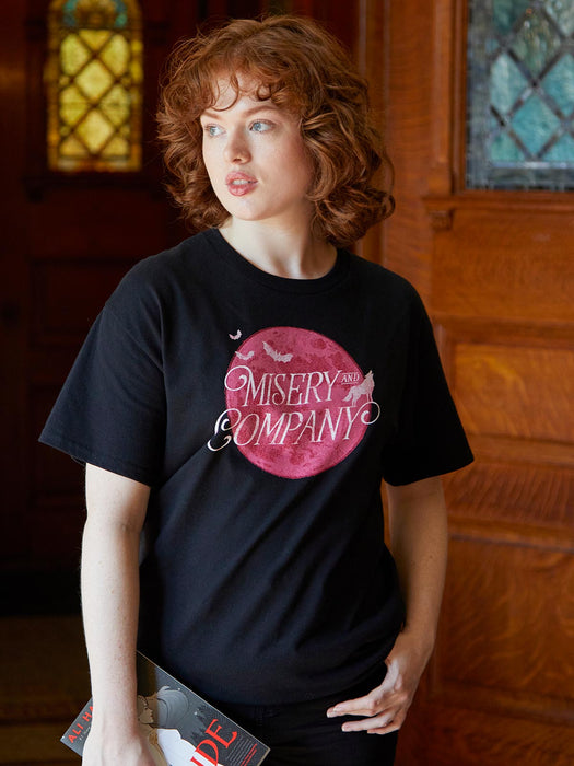 Person wearing a black t-shirt with 'Misery and Company' design, standing indoors.