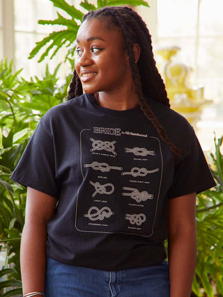 Person wearing a black t-shirt with knot designs and text, standing in a plant-filled room.