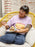 A person sitting in a yellow woven chair wearing a light purple graphic t-shirt with a birthday cake design, blue sweatpants, and holding a plate with a slice of cake.