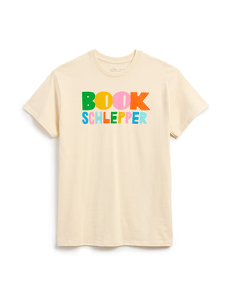 A light beige crewneck t-shirt featuring a colorful graphic print on the chest that reads 'BOOK SCHLEPPER' in playful, multicolored letters, displayed against a clean white background.
