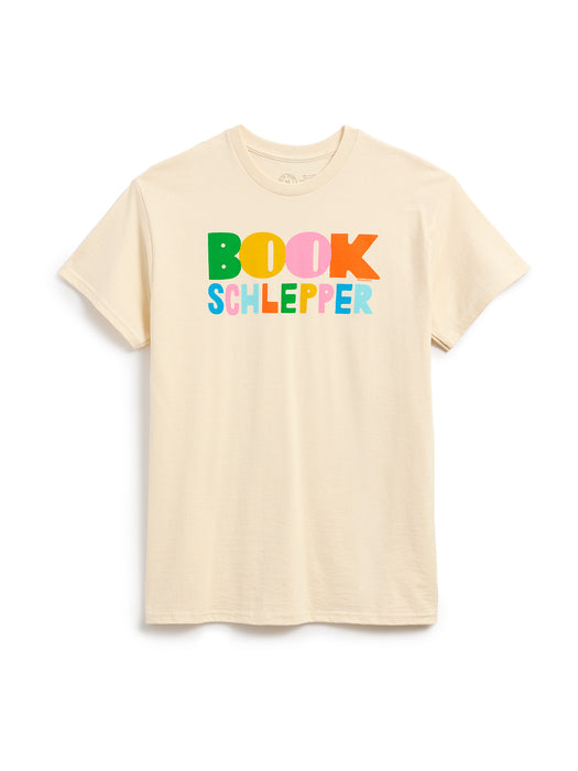 A light beige crewneck t-shirt featuring a colorful graphic print on the chest that reads 'BOOK SCHLEPPER' in playful, multicolored letters, displayed against a clean white background.
