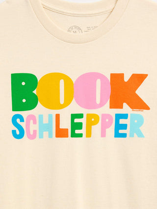 A close-up of a cream-colored t-shirt featuring a colorful graphic print that reads 'BOOK SCHLEPPER' in bold, multicolored letters (green, yellow, pink, orange, blue, and light blue).