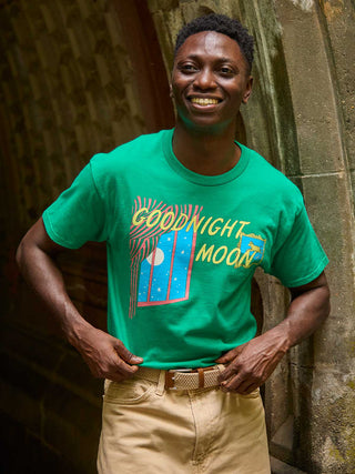 A smiling man wearing a bright green graphic t-shirt with 'GOODNIGHT MOON' and a moon-and-stars design, paired with khaki pants, standing against a stone archway.