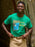 A smiling man wearing a bright green graphic t-shirt with 'GOODNIGHT MOON' and a moon-and-stars design, paired with khaki pants, standing against a stone archway.
