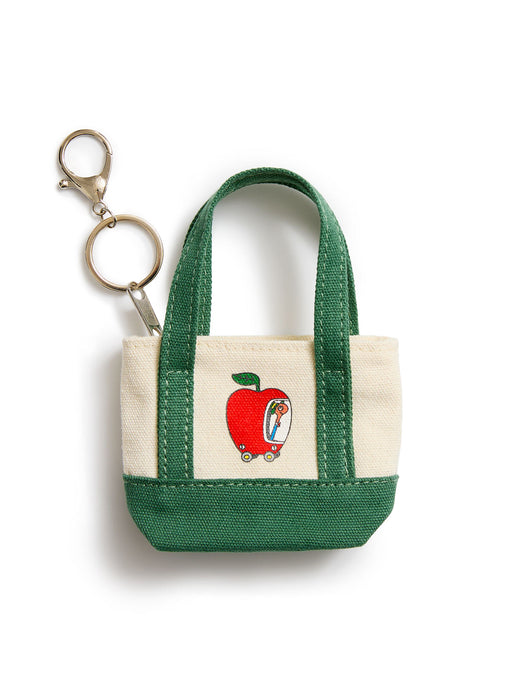 A small canvas tote keychain bag with dark green handles and bottom trim, cream canvas sides, and a red apple graphic on the front, attached to a silver metal key ring on a white background.