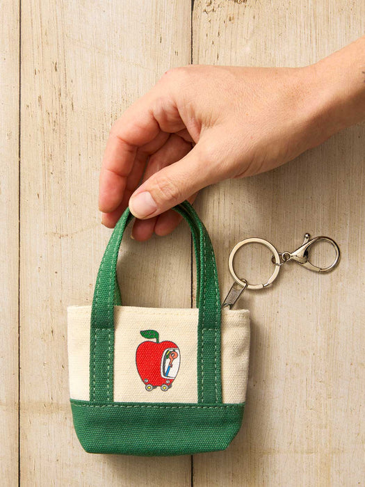 A hand holding a small canvas tote keychain bag with green handles and a cream front panel featuring a red apple illustration, shown with a metal key ring on a light wood background.