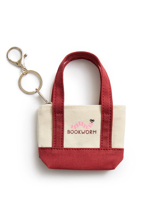 A small canvas tote keychain bag with cream-colored fabric sides, a deep red handle and bottom base, and a gold-tone keyring attached to the side. The front features a cream patch with the brand name 'BOOKWORM' in red and a small worm graphic.