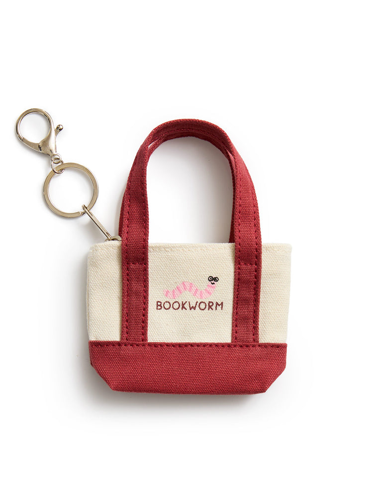 A small canvas tote keychain bag with cream-colored fabric sides, a deep red handle and bottom base, and a gold-tone keyring attached to the side. The front features a cream patch with the brand name 'BOOKWORM' in red and a small worm graphic.