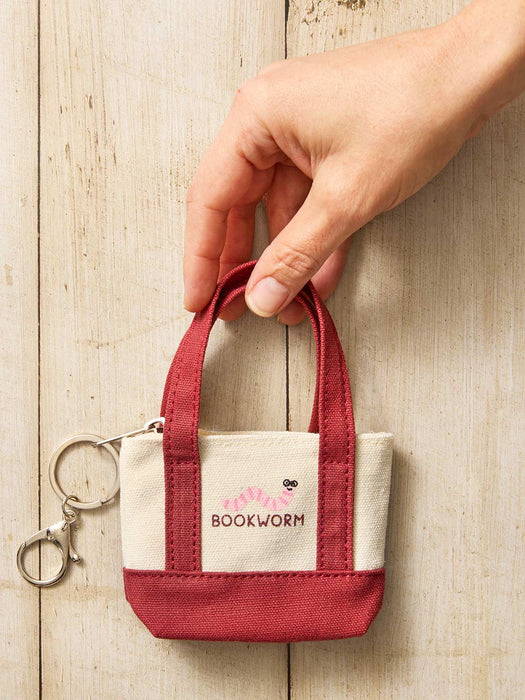 A hand holding a small canvas tote keychain bag with a silver keyring, featuring a cream front panel with a pink worm illustration and the brand name 'BOOKWORM' in dark pink, set against a light wood background.