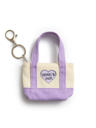 A small canvas tote keychain in lavender and cream with a heart-shaped patch that reads 'ENEMIES TO LOVERS'. The keychain includes a metal split ring and key clasp, photographed on a clean white background.