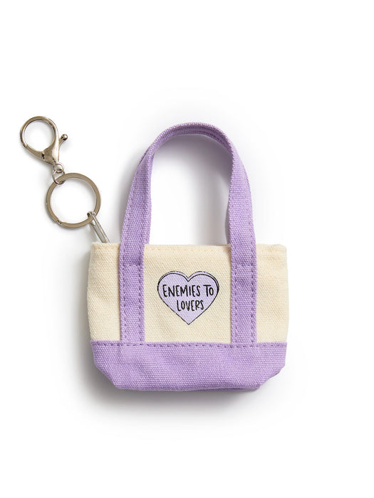 A small canvas tote keychain in lavender and cream with a heart-shaped patch that reads 'ENEMIES TO LOVERS'. The keychain includes a metal split ring and key clasp, photographed on a clean white background.