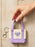 A hand holding a small canvas tote keychain with lavender purple handles and bottom, and a cream center panel featuring a purple heart that reads 'ENEMIES TO LOVERS'. The keychain has a silver lobster clasp.
