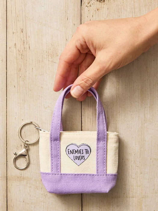 A hand holding a small canvas tote keychain with lavender purple handles and bottom, and a cream center panel featuring a purple heart that reads 'ENEMIES TO LOVERS'. The keychain has a silver lobster clasp.