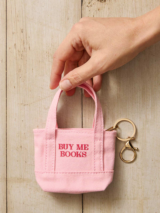 A hand holds a small pink canvas book bag keychain with the words 'BUY ME BOOKS' in red, attached to a gold key ring, against a light wood plank background.