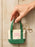 A hand holding a small canvas tote keychain bag with green handles and bottom trim, a cream center panel, pink text reading 'THE BOOK WAS BETTER', and a gold-tone keyring on a light wood plank background.