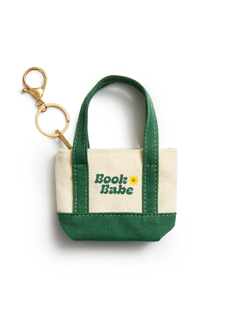 A small canvas tote keychain bag with dark green handles and bottom trim, cream canvas sides, and a gold-tone key ring and clasp. The front is printed with the words 'Book Babe' in green and a small yellow flower.