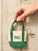 A hand holding a small canvas tote keychain bag with green handles and trim, cream canvas body, 'Book Babe' text on the front, and a gold key ring, set against a light wood plank background.