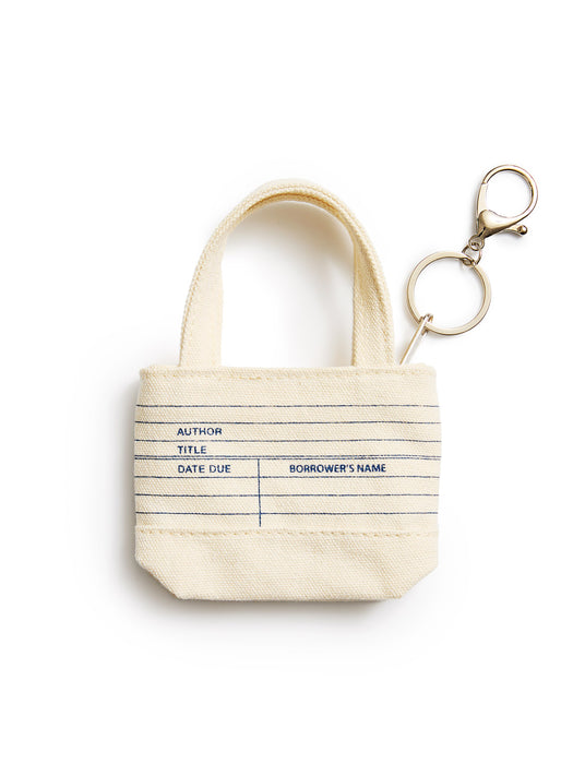 A small canvas tote keychain in cream with blue printed lines and text fields, attached to a gold metal key ring on a white background.