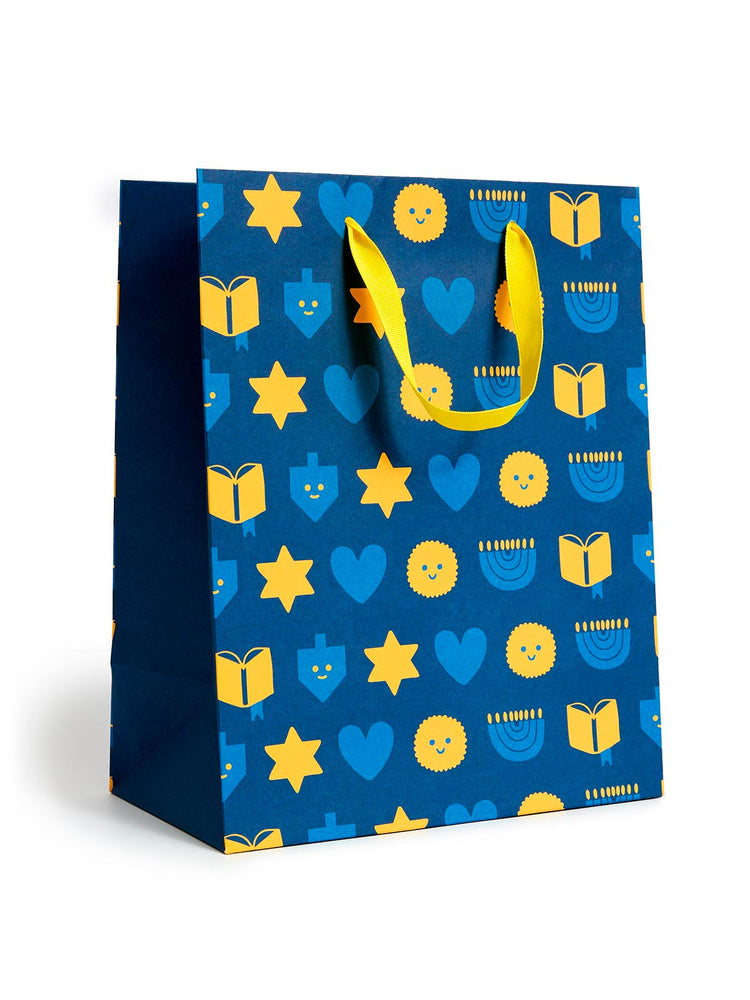 A blue gift bag with a festive pattern of yellow stars, dreidels, menorahs, and open books, featuring bright yellow handles, set against a plain white background.