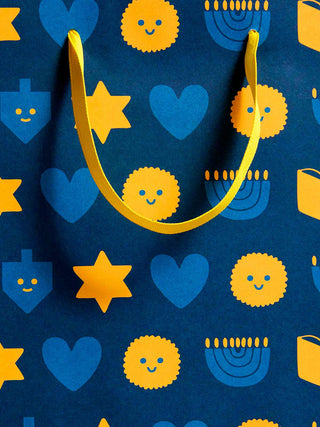 A close-up of a dark blue patterned surface with yellow ribbon handles and repeated yellow and blue holiday-style icons, including stars, hearts, dreidels, and menorahs.