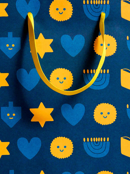A close-up of a dark blue patterned surface with yellow ribbon handles and repeated yellow and blue holiday-style icons, including stars, hearts, dreidels, and menorahs.