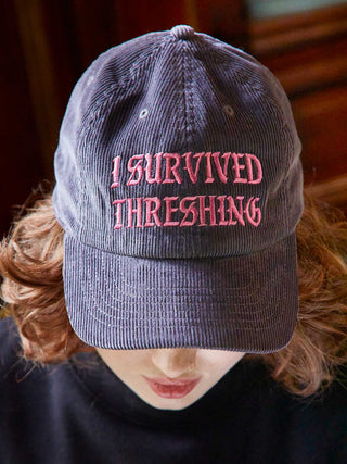 Gray cap with 'I Survived Threshing' text worn by a person with red hair.