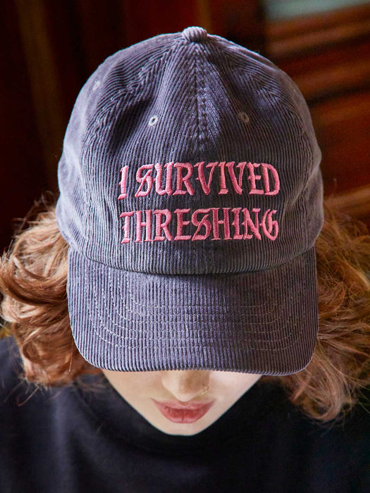 Gray cap with 'I Survived Threshing' text worn by a person with red hair.