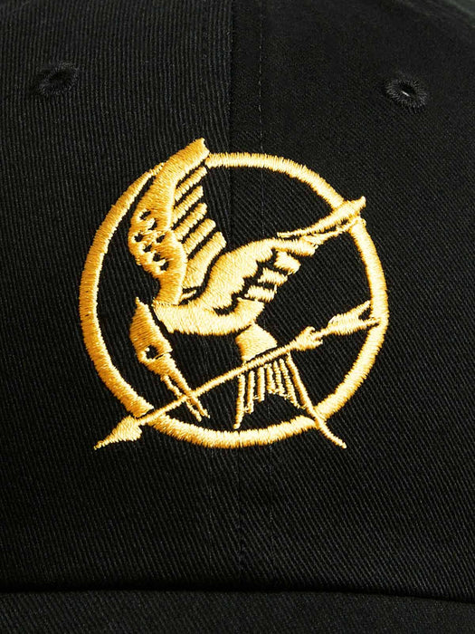 Close-up of a black baseball cap featuring a gold embroidered Hunger Games Mockingjay logo.