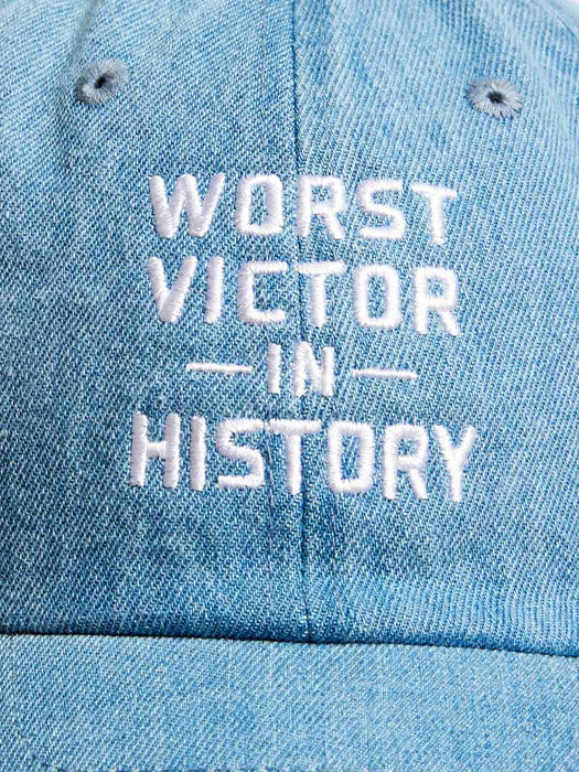 A close-up of a light blue denim baseball cap featuring white embroidered text that reads 'WORST VICTOR IN HISTORY'.