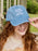 A smiling woman with long red hair wears a light blue denim baseball cap with white embroidered text reading 'WORST VICTOR IN HISTORY'. She is outdoors on a porch with greenery in the background.