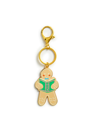 A gingerbread man keychain charm with a gold-tone metal finish, featuring a green book and small red and blue facial details, attached to a gold split ring and lobster clasp on a white background.