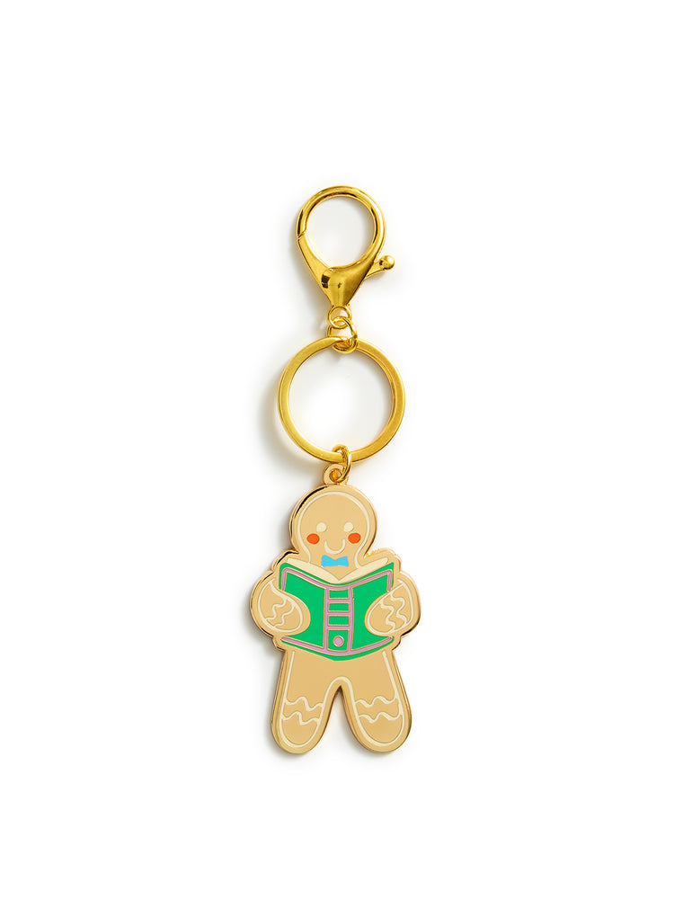 A gingerbread man keychain charm with a gold-tone metal finish, featuring a green book and small red and blue facial details, attached to a gold split ring and lobster clasp on a white background.