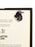 A dragon-shaped bookmark with a pink border and black, gray, and yellow illustration, attached to a printed page inside a black book.