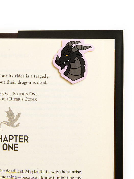 A dragon-shaped bookmark with a pink border and black, gray, and yellow illustration, attached to a printed page inside a black book.
