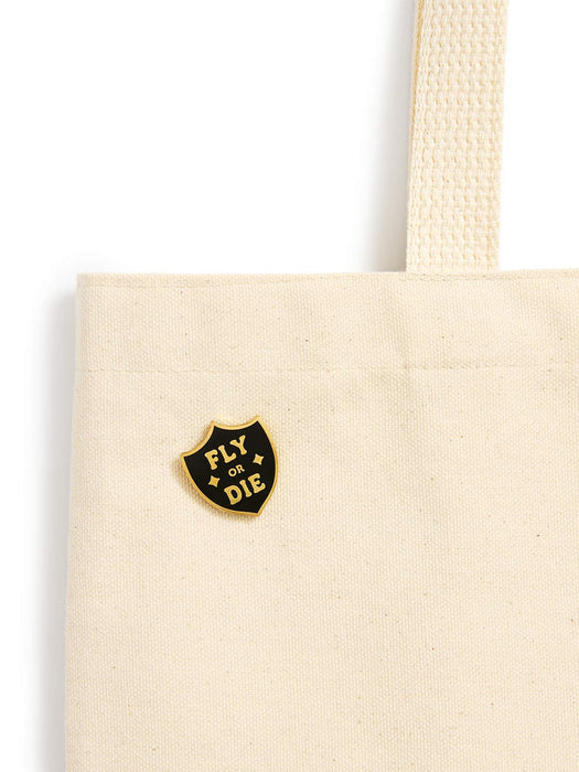 A close-up of a cream canvas tote bag with a black and gold shield-shaped enamel pin that reads 'FLY OR DIE' on the front, set against a plain white background.