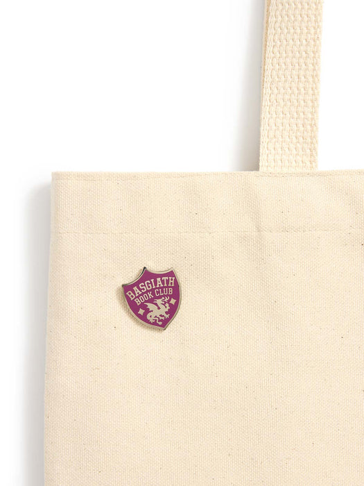 A close-up of a cream canvas tote bag with a small purple and gold Basgiath Iron Club shield-shaped pin on the front, set against a plain white background.