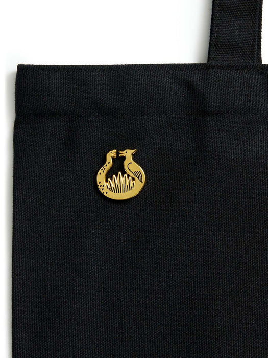 A close-up of a black canvas tote bag featuring a small gold sunrise on the reaping enamel pin on the front, set against a plain white background.