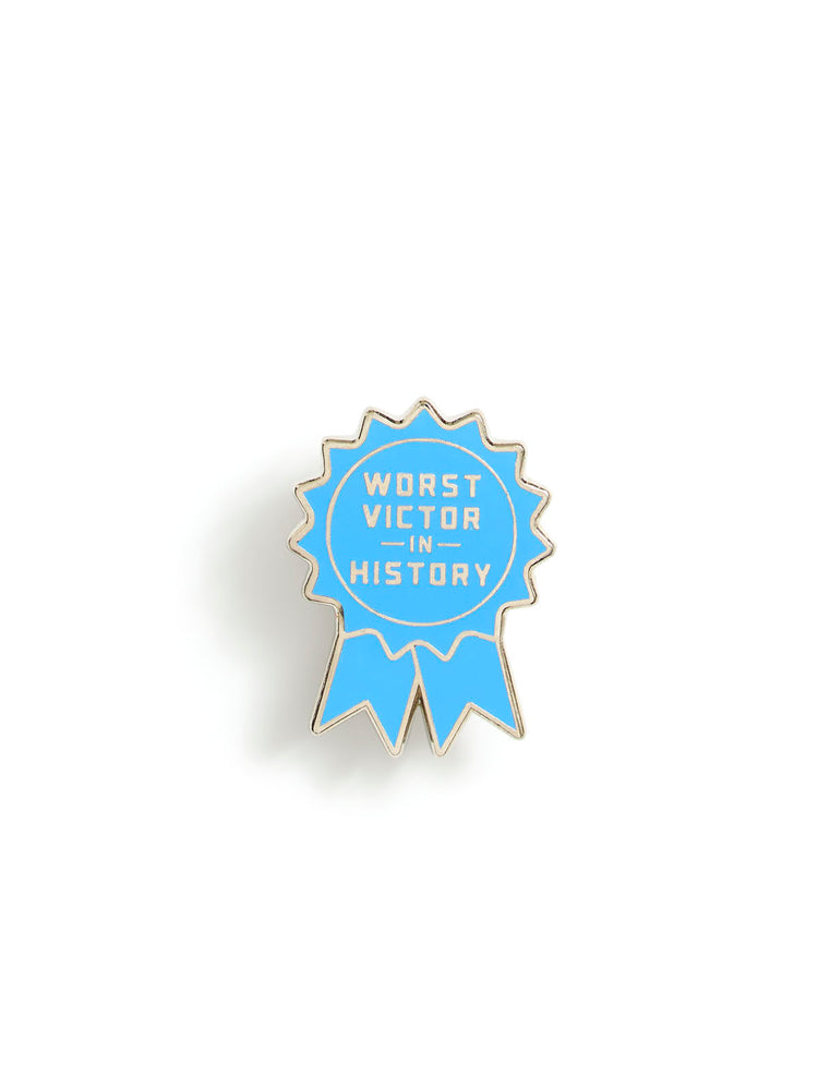 A light blue ribbon-shaped enamel pin with gold metal edging and the text 'WORST VICTOR IN HISTORY' in white, shown on a plain white background.
