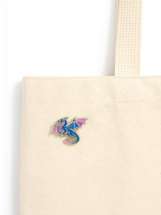 A close-up of a cream canvas tote bag with a small colorful dragon enamel pin on the front, set against a plain white background.