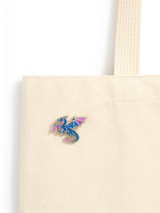 A close-up of a cream canvas tote bag with a small colorful dragon enamel pin on the front, set against a plain white background.