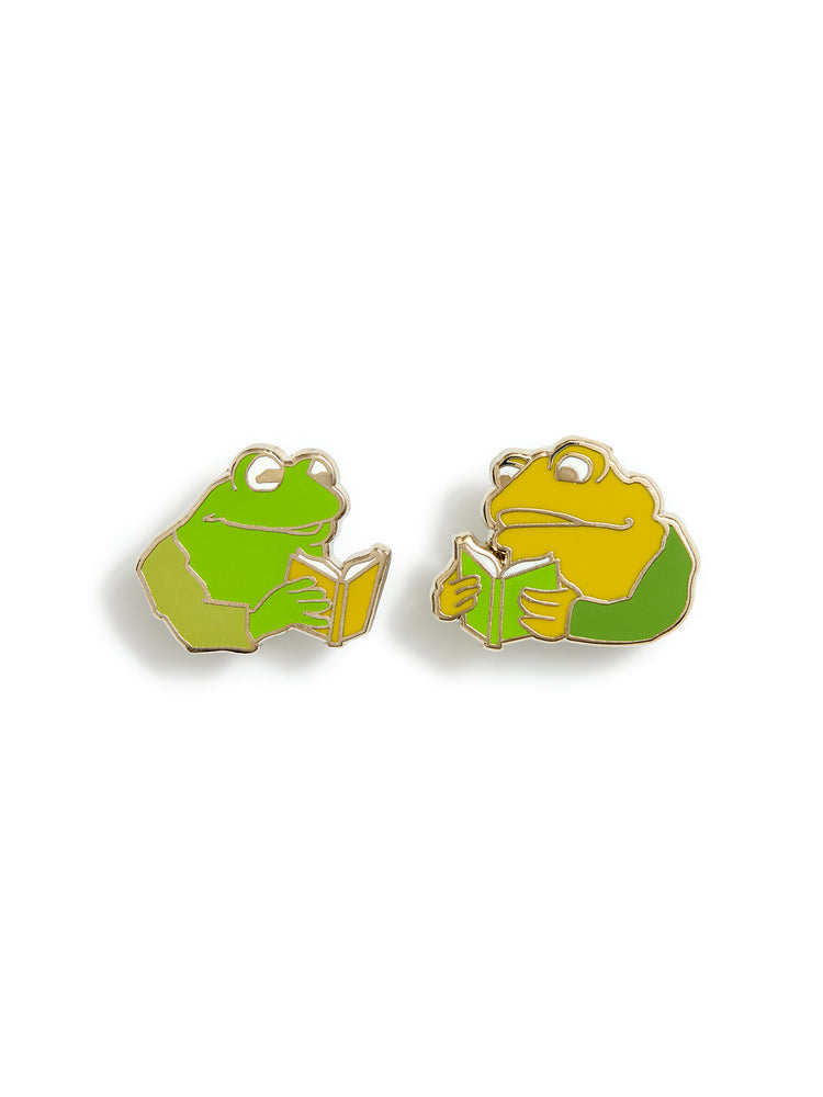 Two frog and toad shaped enamel pins on a white background, one green and one yellow, both holding and reading a small book.