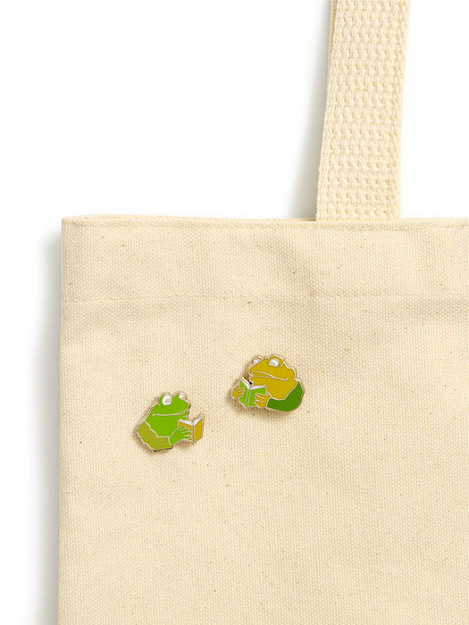 Two small frog and toad shaped enamel pins in green and yellow are shown on a beige canvas tote bag against a white background.