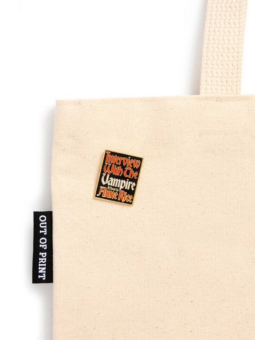 A close-up of a beige canvas tote bag with a black fabric brand tag reading 'OUT OF PRINT' and a small square enamel pin that says 'Interview With The Vampire Bride Price' in red and white text.