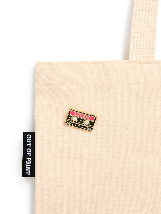 A close-up of a beige canvas tote bag with a small cassette tape enamel pin on the side. The pin has pink, gold, and black details.