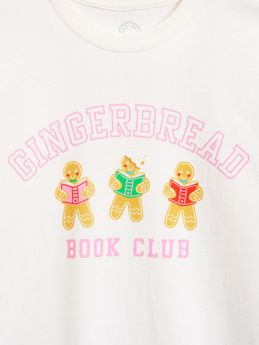 A close-up of a white crewneck t-shirt featuring a colorful graphic print. The design displays the words 'GINGERBREAD BOOK CLUB' in pink lettering, with three gingerbread characters reading books in the center.