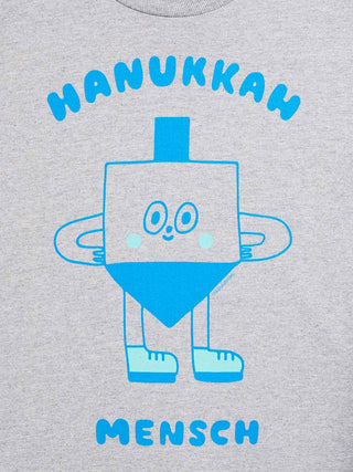 A close-up of a light heather gray t-shirt featuring a blue graphic of a dreidel character with the text 'HANUKKAH' above and 'MENSCH' below in blue.